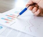 A close-up of a hand with a pen analyzing data on colorful bar and line charts on paper.